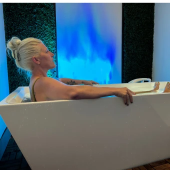 Person resting their arms on the sides of a white cold-plunge tub beside a blue illuminated wall.