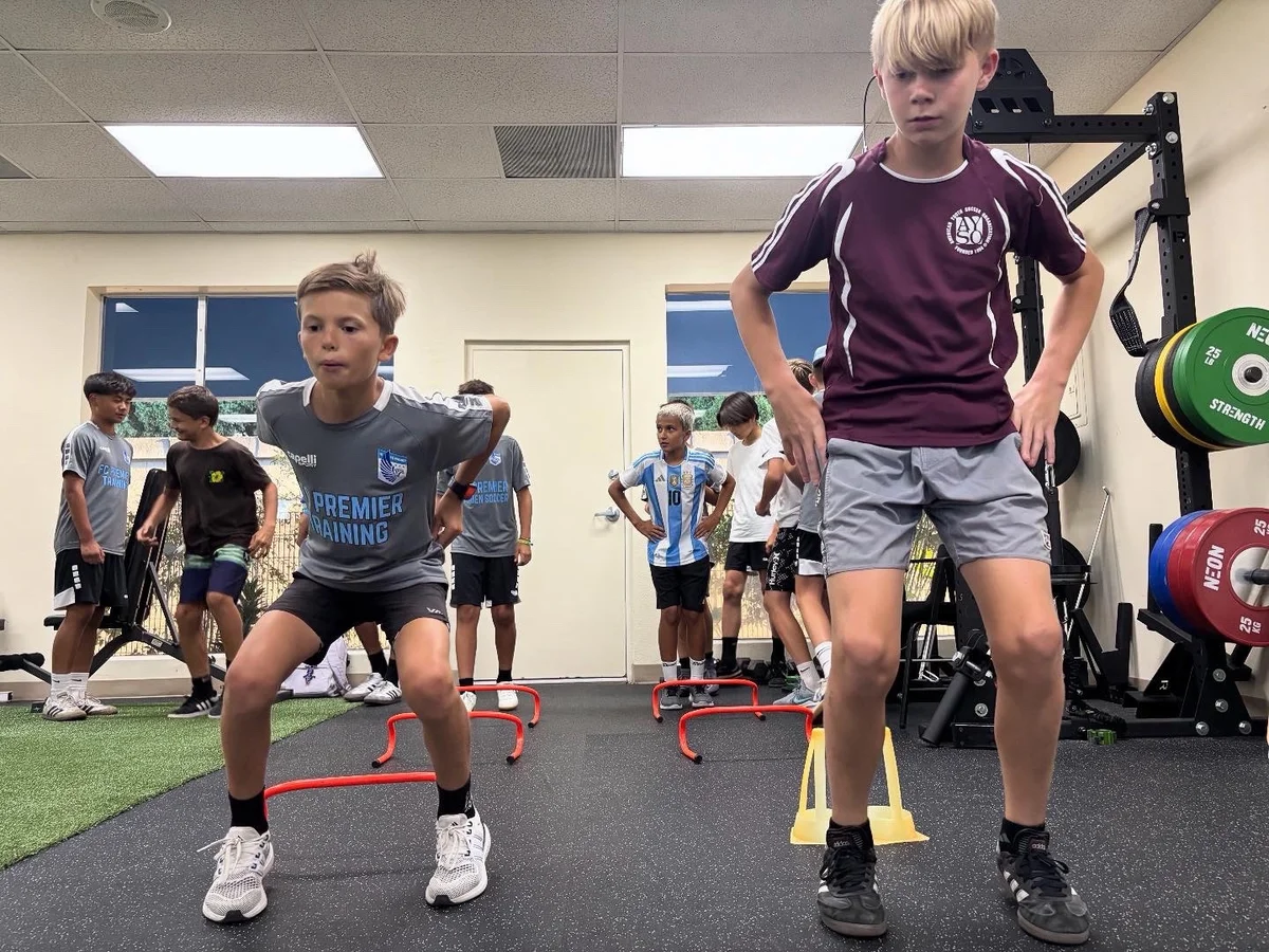 Young athletes practicing agility drills with small hurdles in an indoor training space.